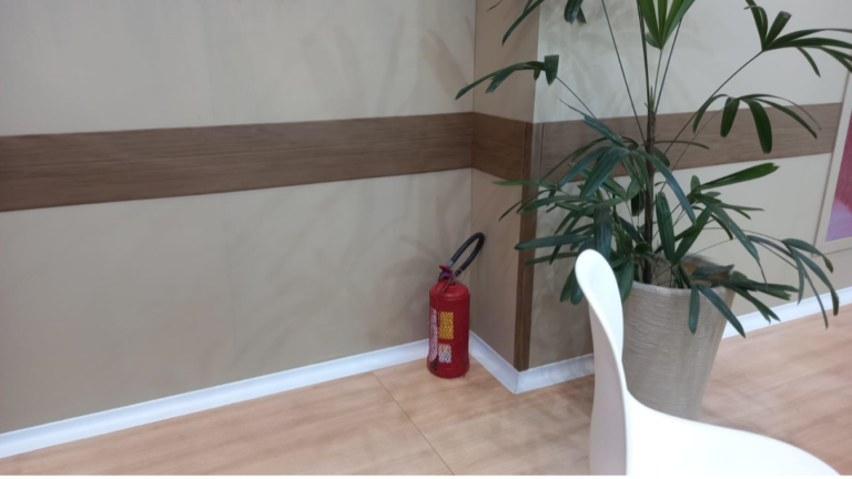 Protetor de parede amadeirado instalado em ambiente interno com planta decorativa e extintor ao fundo.