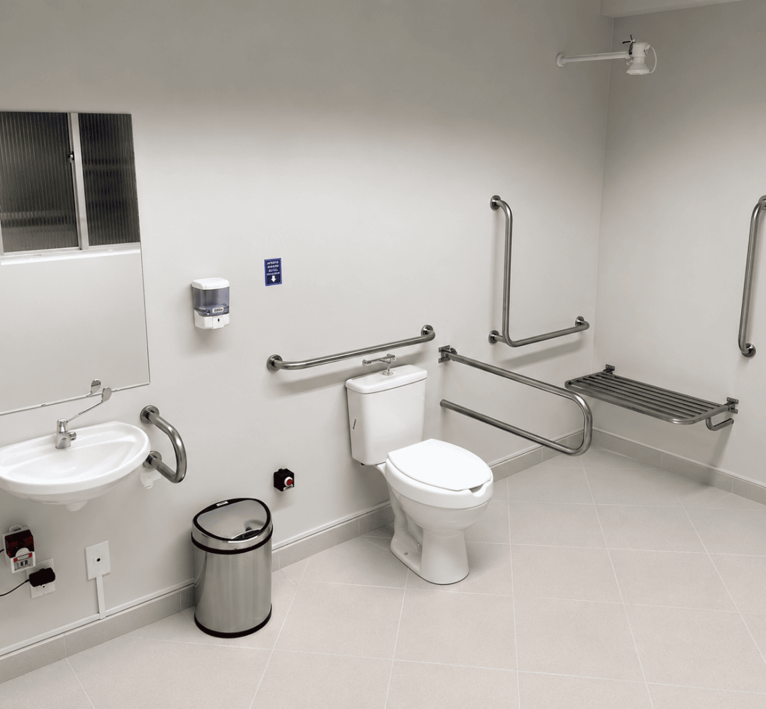 Banheiro acessível com barras de apoio em inox instaladas ao redor do vaso sanitário e da pia, incluindo barras laterais, barras de canto e banco articulado para apoio. O ambiente possui acabamento claro, espelho, dispenser de papel e espaço amplo para circulação de pessoas com mobilidade reduzida.
